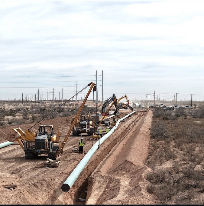 A pipeline construction site in an arid landscape with construction equipment, including side-boom tractors, and workers laying a large pipe in a trench.