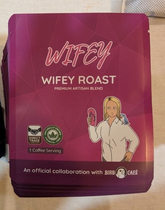 A purple coffee packet labeled 'WIFEY WIFEY ROAST PREMIUM ARTISAN BLEND' featuring a cartoon blonde woman holding a pink object with a blue parrot, marked as a collaboration with Birb Café, Direct Trade, and Ethically Grown.