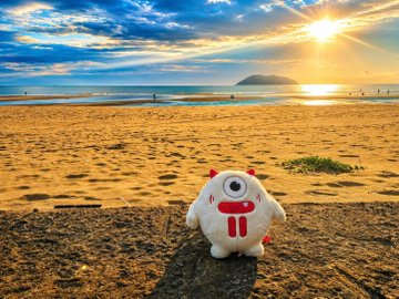 A small white plush mascot with red horns and a red 'T' logo sitting on sandy rocks at a beach during golden hour, with the sun setting over the ocean.