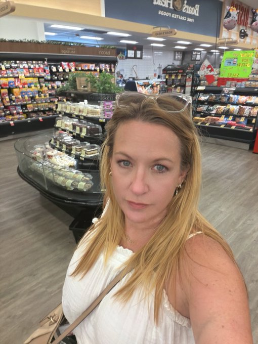 A blonde woman wearing a white top and sunglasses perched on her head takes a selfie inside a grocery store, with a floral display and "Fresh" signage visible in the background.