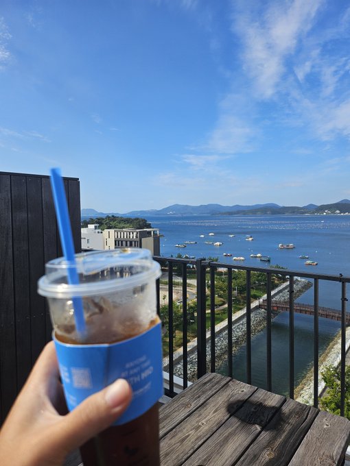 A hand grasps a plastic cup with a blue straw containing an iced beverage, with a scenic view of a bridge, water, boats, and mountains in the background under a blue sky.