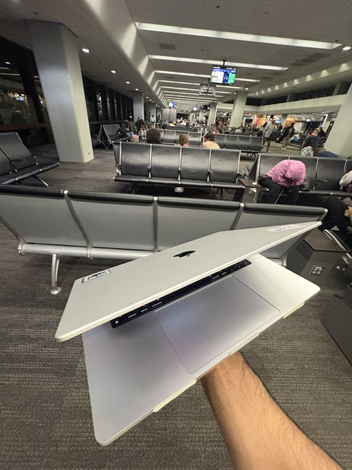 The image shows a person holding an open laptop computer with a sticker on its back. The laptop appears to be a Macbook Pro, which has a silver color scheme. In the background of the image, there are several people sitting or standing around in what seems to be an airport terminal waiting area.