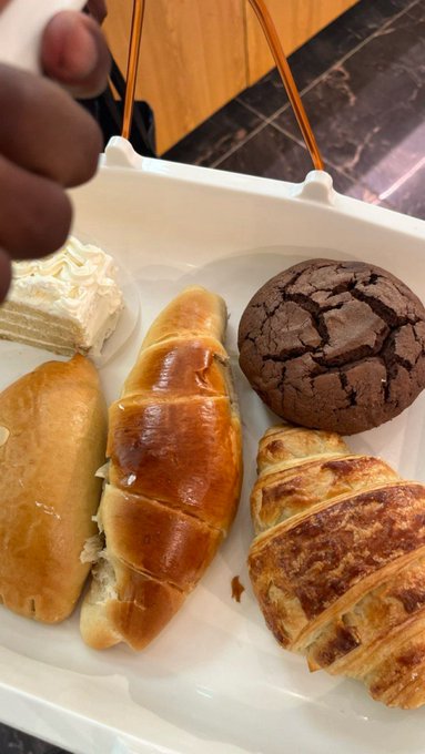 A white container holds an assortment of pastries including a slice of layered cake, bread rolls, a chocolate cookie, and a croissant.