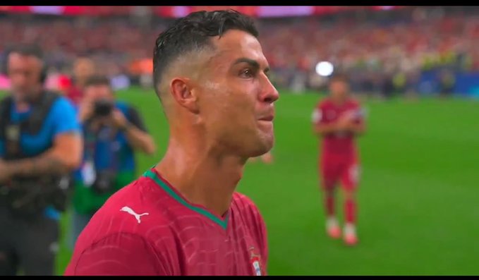 Cristiano Ronaldo in a red Portugal national team jersey (with Puma logo) on a soccer pitch, appearing emotional with photographers and other players visible in the background.