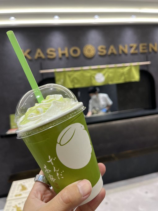 Hand holding a green matcha drink in a clear cup with a green straw, with a 'KASHO SANZEN' storefront visible in the background.