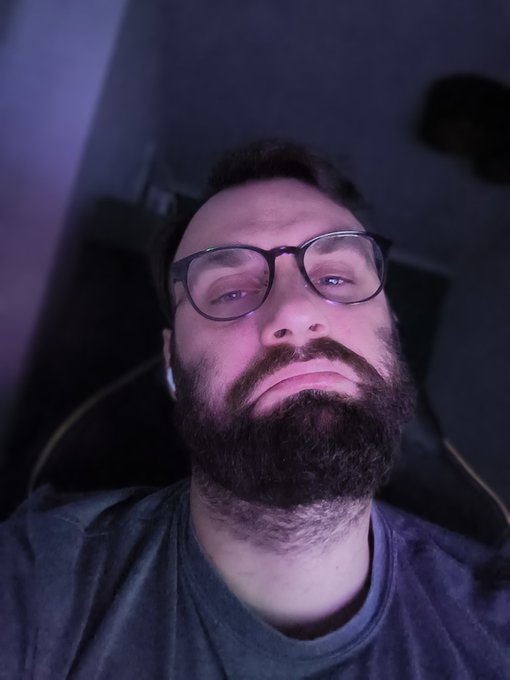 The image features a man with glasses, a beard, and a mustache wearing a gray shirt. He has his eyes closed and appears to be looking at the camera directly. The background of the photo shows a purple wall, which contrasts with the man's dark hair color. There is also an earphone visible in the scene, suggesting that he might have been listening to music or a podcast before taking the picture.