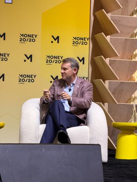 A man speaks while seated on a stage at Money 2020, backed by repeated “M” logos and the text “MONEY 20/20.” The conference setting and visible branding reinforce the post’s discussion of an interoperable, hybrid future for public and private finance networks.