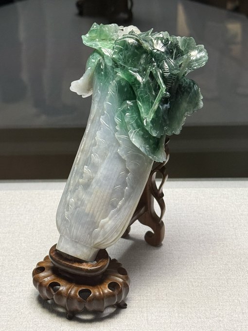 A polished jade sculpture of a leafy vegetable, likely Chinese cabbage, displayed on a carved wooden stand in what appears to be a museum setting.