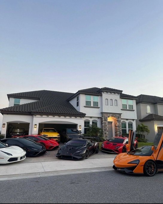 A luxury home with a tile roof has an impressive collection of high-end sports cars parked in the driveway, including Lamborghinis, Ferraris, and McLarens.