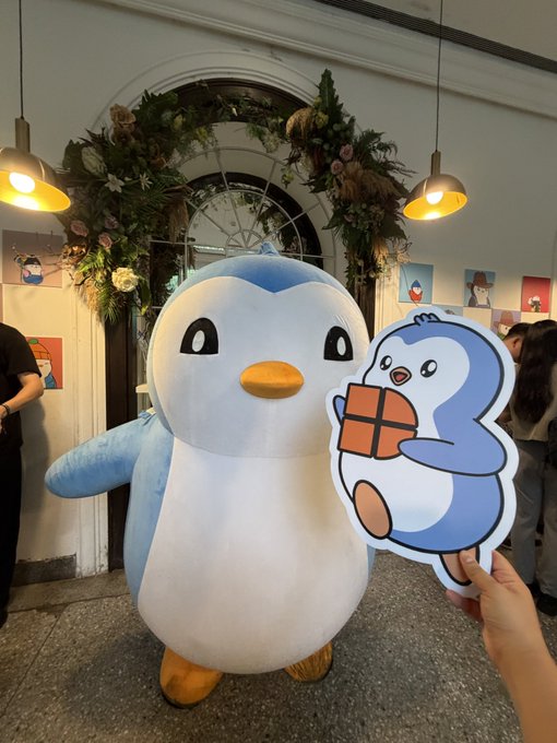 A person holds a cardboard cutout of the character Piplup in front of a person in a Piplup mascot costume.