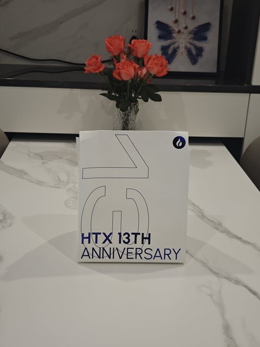 A white display card on a marble table features a large "13" design and reads "HTX 13TH ANNIVERSARY," with a vase of orange roses in the background.