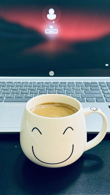 A white smiley-face coffee mug filled with espresso, sitting in front of an open HP laptop displaying a user profile icon.
