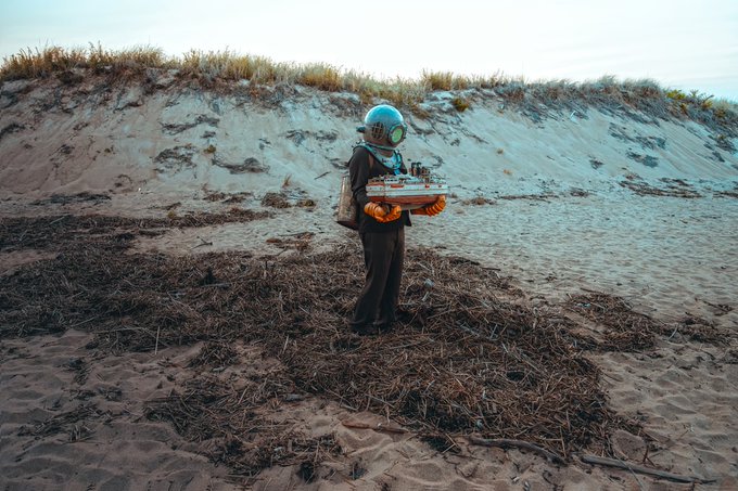 A person in a vintage diving suit and helmet stands on a sandy beach holding a case, with grassy dunes and the ocean in the background.