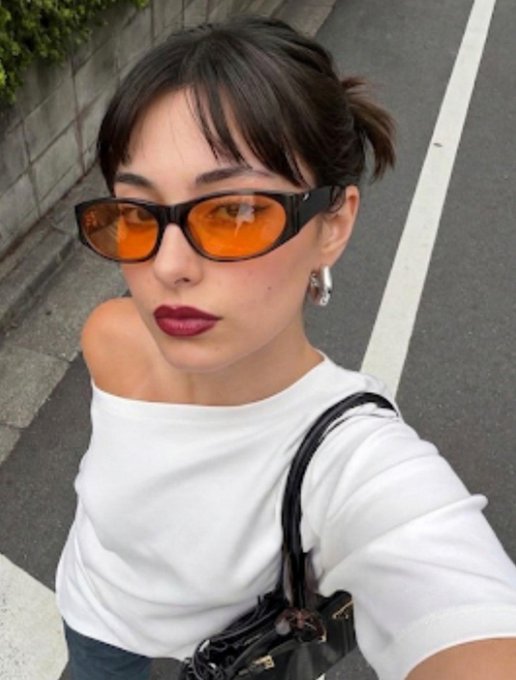 A selfie of a stylish woman wearing orange-tinted sunglasses, dark lipstick, a white off-shoulder top, and carrying a black handbag, standing on a street.