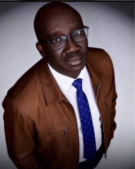 Portrait of a man wearing glasses, a brown leather jacket, white shirt, and a blue patterned tie.