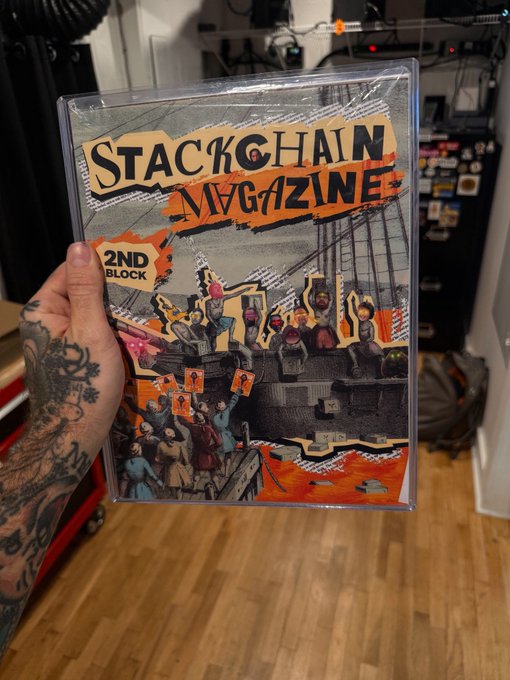 A tattooed hand holding up a plastic-wrapped copy of 'STACKCHAIN MAGAZINE 2ND BLOCK', which features a classical painting-style cover with people protesting and holding signs with the Bitcoin '₿' logo.