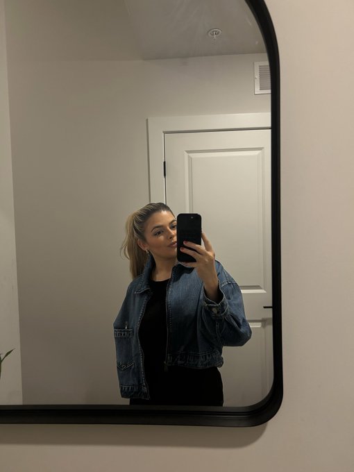 A mirror selfie of a woman with blonde hair wearing a blue denim jacket over a black top, holding a smartphone in a room with a white door and vent visible in the reflection.