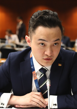The image shows a young Asian man in a dark navy suit, white shirt, and striped tie, seated at what appears to be a formal conference or meeting setting with a lanyard badge around his neck.

The caption likely relates to the accompanying post about Justin Sun, the founder of TRON cryptocurrency, who is pictured here as the central figure in the lawsuit dispute with World Liberty Financial (WLFI) over frozen user assets.