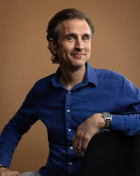 A confident man in a blue patterned button-up shirt smiles warmly at the camera against a warm brown backdrop, though no text, names, or identifying details are visible in the image. The portrait does not appear to directly relate to the post about Zcash's shielded pools and cryptographic proof developments, as the subject's identity or connection to the cryptocurrency project cannot be determined from the visual alone.