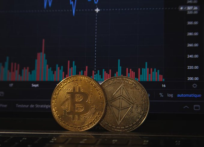 Physical Bitcoin and Ethereum coins sit in front of a laptop screen displaying a cryptocurrency price chart with red and green candlesticks, showing prices ranging from $200 to $340.
