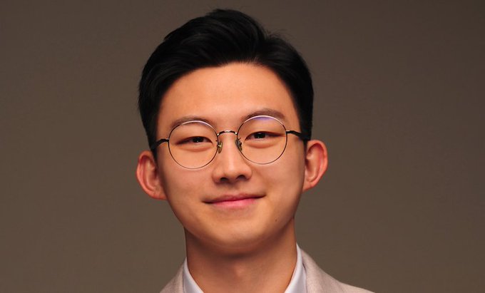 A young East Asian man with short black hair and round wire-rimmed glasses smiles softly at the camera while wearing a light-colored blazer over a white shirt. The post's note about Bitcoin's bull market confirmation appears unrelated to the portrait image.