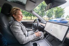 A woman sitting in the driver's seat of a modern vehicle, looking at a large touchscreen display on the dashboard, possibly a Tesla or similar EV.