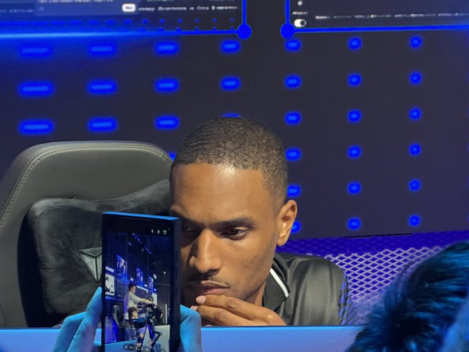 A focused man, presumably the crypto trader known as Ansem, leans forward while filming himself on his phone against a backdrop of a dark screen displaying blue grid patterns and charts. The setup captures his concentrated "locked in" trading session, with the phone screen showing the recording in progress.