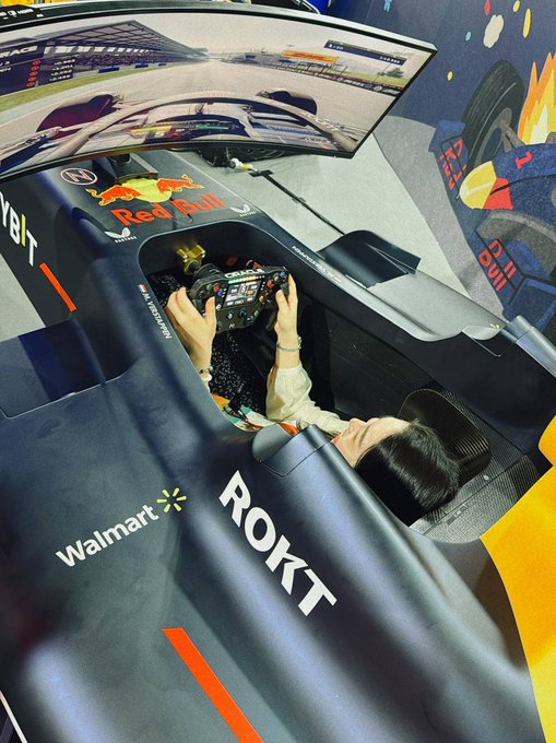 A person sits inside a Formula 1 simulator cockpit branded with Red Bull, Walmart, and ROKT logos, holding a steering wheel in front of a monitor displaying a racing game.