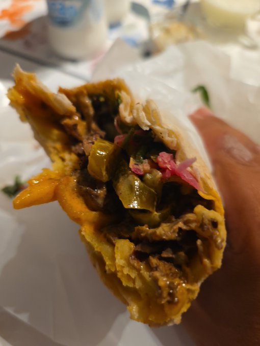 A close-up of a partially eaten wrap or burrito being held, revealing filling of meat, pickled vegetables, and onions, with another sausage visible in the background.