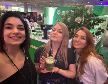 The image shows three women standing together, each holding a drink - one of them has a green smoothie while the other two are holding drinks with white tops. They appear to be posing for a photo at an event or gathering. The background includes a display table and some potted plants, suggesting that they might be attending a social function or a similar occasion.