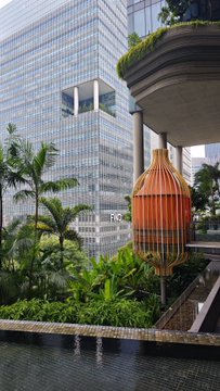 An architectural view showing a modern glass office building behind a decorative garden with tropical plants, palm trees, and an ornate hanging lantern-style sculpture over a water feature.