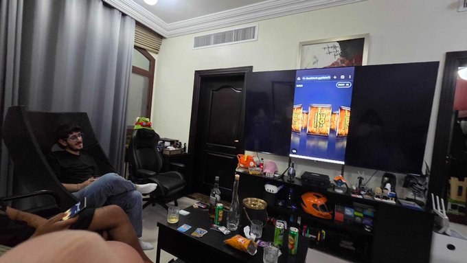 A casual living room scene where a man with glasses sits on a couch watching TV, which displays beverage cans, while another person holds a phone in the foreground.
