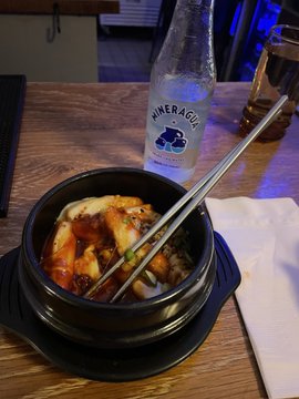 A bowl of Korean bibimbap-style rice dish in a stone pot with metal chopsticks, next to a bottle of Mineragua sparkling water on a wooden table.
