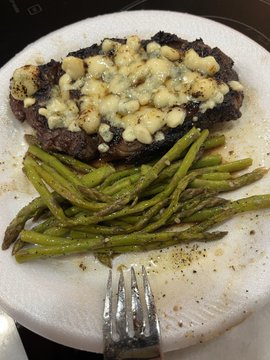 A white plate containing a steak topped with melted blue cheese alongside a portion of cooked green beans, with a silver fork resting at the bottom of the plate.