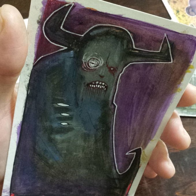 A hand holding a dark, hand-painted artwork of a horned, monstrous figure with glowing eyes, set against a purple background.