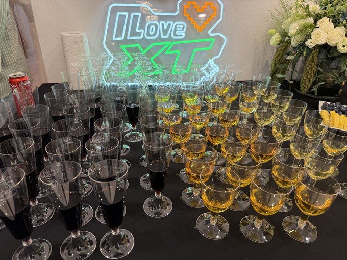 A lavish drink station features rows of champagne flutes filled with white wine and dark beverages, set against a glowing neon "I Love XT" sign celebrating the 8th anniversary of XT. Elegant floral arrangements and a fruit platter complete the festive setup, fitting the celebratory mood of the company milestone.