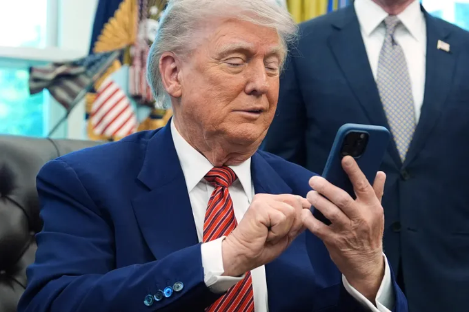 A photograph of Donald Trump in a blue suit and red striped tie looking at and pointing at a blue smartphone, with another person in a suit standing behind him near American flags.