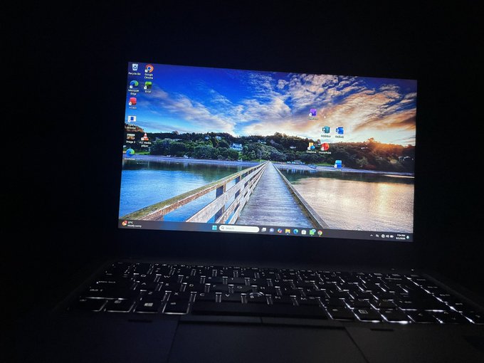 A dark photo of an open laptop displaying a scenic landscape wallpaper of a wooden pier extending into a calm lake at sunset.
