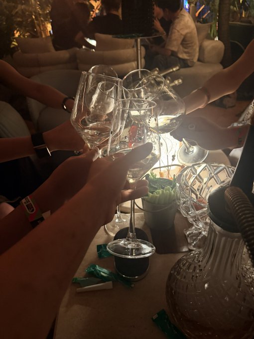 Multiple hands clink wine glasses together in a toast at a dimly lit lounge or club, with a glowing table centerpiece, crystal glassware, and a hookah base visible in the foreground.