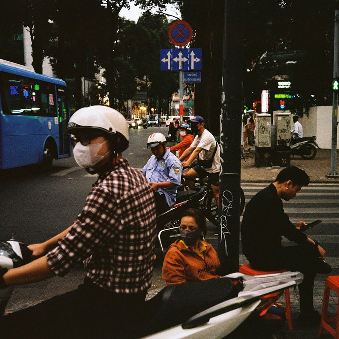 A street scene in Vietnam showing a woman in a plaid shirt and helmet on a motorbike, with a police officer, pedestrians, a blue bus, and street signs including a no-parking sign.