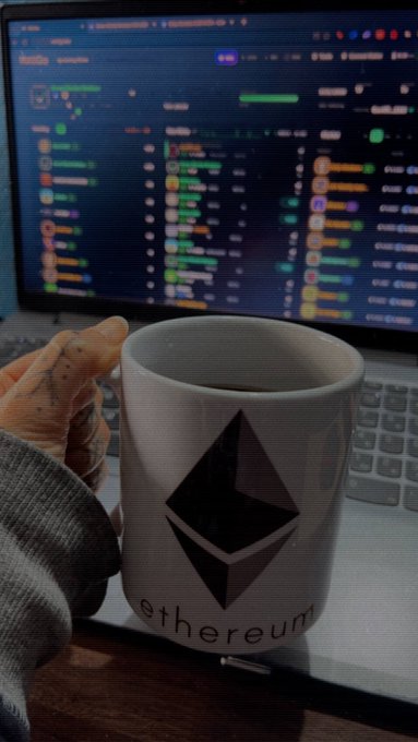 A hand holds a white ceramic mug featuring the Ethereum logo and 'ethereum' text, with a computer monitor displaying a cryptocurrency trading chart in the background.