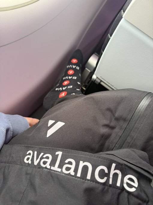 The image shows a person's feet resting on a black backpack with white text, which reads "avax team 1", suggesting that they are traveling or embarking on an adventure. The backpack appears to be filled with various items, indicating that the traveler is prepared for their journey.