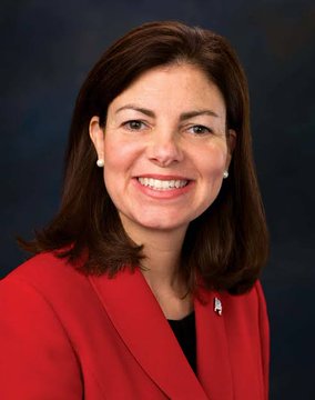 Kelly Ayotte, Governor of New Hampshire, is pictured in a professional headshot wearing a red blazer and a small American flag pin. Her image accompanies a post highlighting her public support for considering Bitcoin adoption at the state level.