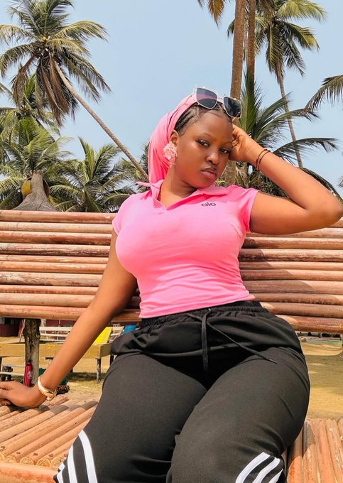 A woman wearing a pink polo shirt, black sweatpants with white stripes, and a pink headscarf sits on a bamboo bench with palm trees and a beach in the background.