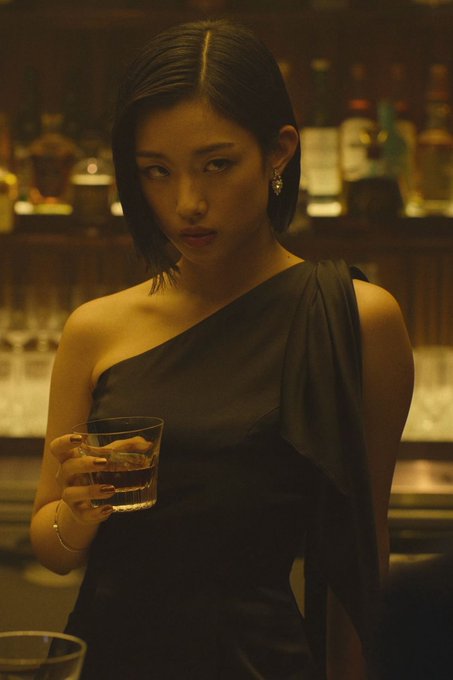 A woman in a black one-shoulder dress stands at a warmly lit bar holding a drink, with bottles blurred in the background.