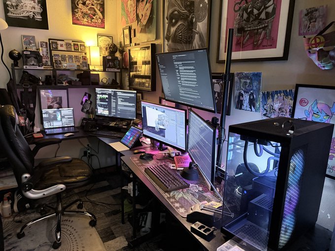 A multi-monitor computer setup is surrounded by various posters and framed artwork in a dimly lit room.