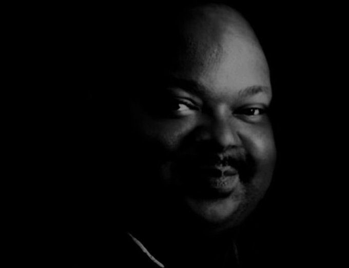 A dramatic black and white close-up portrait of a bald man with a mustache, partially illuminated against a dark background.