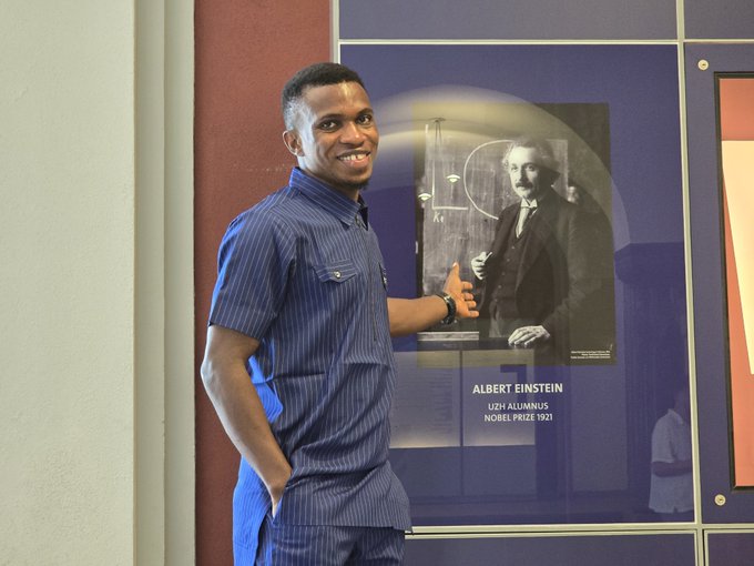 A man in a blue striped shirt smiles and gestures toward an Albert Einstein poster labeled 'ALBERT EINSTEIN, UZH ALUMNUS, NOBEL PRIZE 1921'.