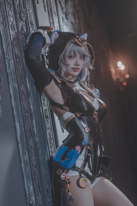A cosplayer dressed as "Cipher," a character with silver hair and a hooded outfit featuring intricate black and gold detailing, poses confidently against an ornate backdrop. Her costume includes distinctive blue accessories and star-shaped embellishments, capturing the futuristic and enigmatic aesthetic associated with the character.
