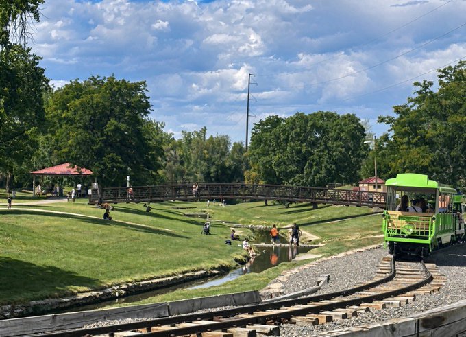 A green miniature park train travels along tracks past a small bridge and pond where visitors enjoy the sunny day, perfectly capturing the "touching some grass" sentiment before heading back to work to "get ppl paid."